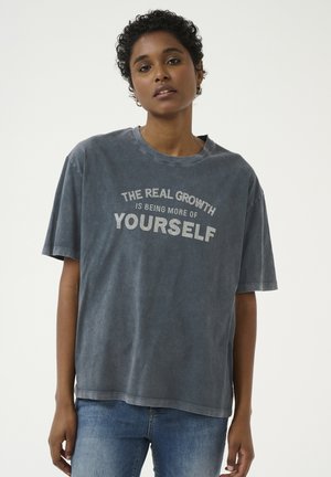 Overdimensioneret grå t-shirt lavet af bomuld, med rund halsudskæring og korte ærmer. Teksten i hvid læser: "DEN VIRKELIGE VÆKST ER AT BLIVE MERE AF DIG SELV."