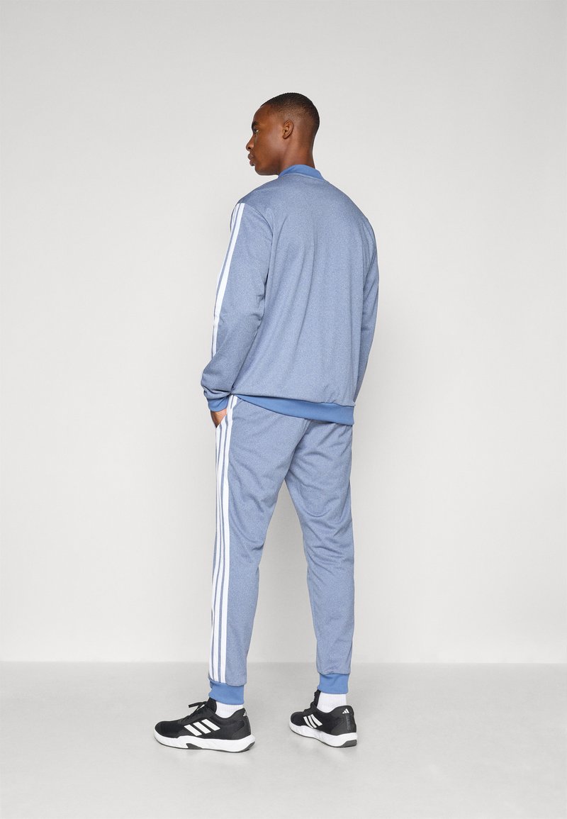 Survêtement bleu clair avec des bandes blanches le long des manches et des côtés, fait d'un tissu lisse et texturé. Comprend une veste à col et un pantalon ajusté.