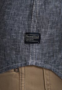 Tessuto a righe blu e bianco con un'etichetta nera che recita "Petrol Ind." visibile; pantaloni marrone chiaro con dettaglio a bottone in fondo.