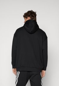 Hoodie preto com acabamento texturizado, design oversized e capuz com cordão. Apresenta punhos e bainha canelados. O modelo usa calças de fato de treino pretas com riscas.