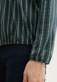 Grüner gemusterter Top mit vertikalen Streifen in Marineblau, Weiß und Hellgrün. Weicher Stoff, geraffte Ärmelbündchen und runder Saum. Denim-Hintergrund.