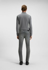 Bărbat întors, purtând un tricou polo gri cu mânecă lungă, blugi gri slim-fit și adidași negri, pe un fundal simplu deschis.