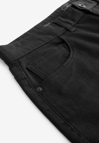 Close-up af sorte denimbukser, der viser forlomme, lille møntlomme, bæltestrop og metalnitte-detalje.