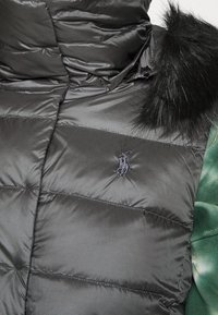 Svart pufferväst med quiltat mönster, med hög krage och en pälsdetalj. Logotyp broderad på vänster sida. Mjuk, glänsande yta.