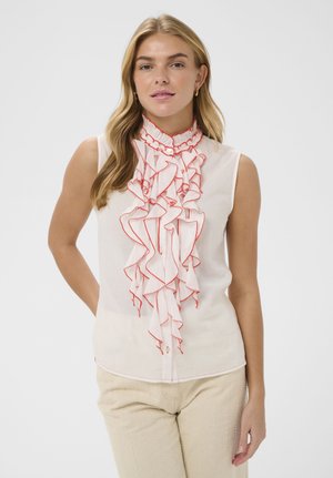 Blonde Frau, die eine ärmellose weiße Bluse mit roten Rüschen entlang der Vorderseite trägt, kombiniert mit hellbeigen Hosen, stehend vor einfarbigem Hintergrund.