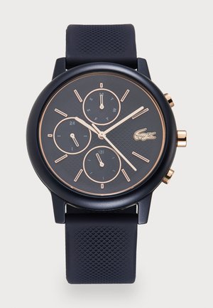 Montre noire avec un cadran texturé, accents en or rose, trois sous-cadrans et un bracelet en silicone. Présente un boîtier rond et une quincaillerie à bouton-poussoir.
