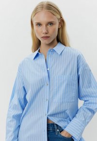 Light blue button-up shirt with white vertical stripes, featuring a chest pocket and a classic collar, made of a smooth, woven material.