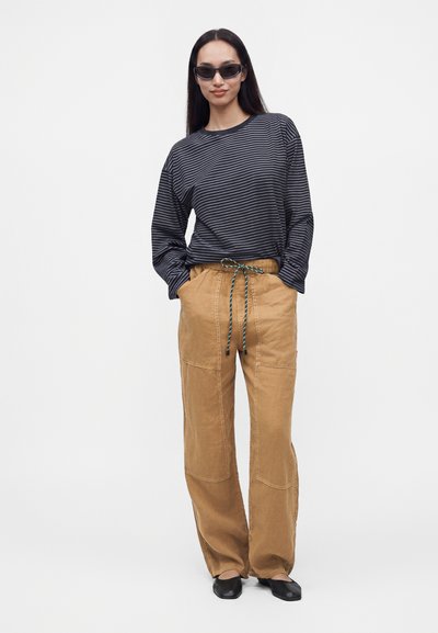 Femme portant une chemise à manches longues rayée foncée, un pantalon beige ample avec un cordon, des chaussures noires et des lunettes de soleil, debout, les mains dans les poches.