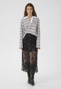 Kvinde iført en grå og hvid stribet langærmet skjorte, sort transparent blomster-lace nederdel og sorte højhælede støvler, stående på hvid baggrund.