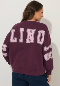 Felpa oversize color marrone scuro con grandi lettere rosa a blocchi "LINO" sul retro e il numero "23" sul braccio sinistro. Materiale in cotone confortevole.