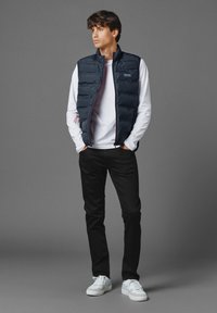 Marinblå vadderad väst med horisontell quiltning, röda accenter, bärs över en vit långärmad skjorta, tillsammans med svarta byxor och vita sneakers.