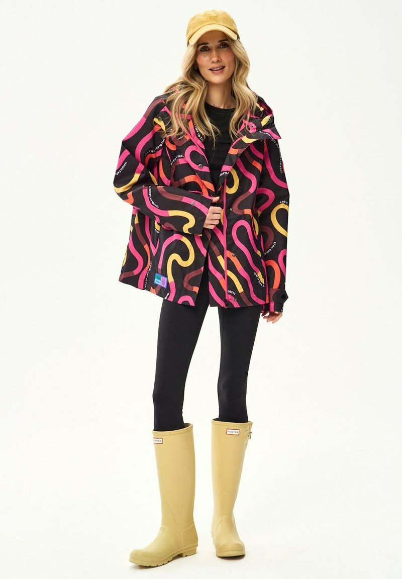 Blondynka w żółtej czapce, czarnych legginsach, kolorowej kurtce z wzorem i beżowych, sięgających kolan kaloszach stojąca na białym tle.