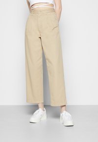 Beige weit geschnittene Hose aus Baumwolle mit hoher Taille, vorderen Falten und zwei Seitentaschen. Kombiniert mit weißen Sneakers.