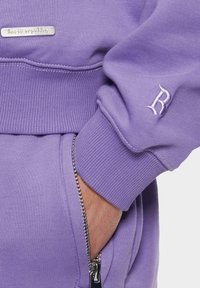 Lavendelfarbenes Sweatshirt mit gerippten Bündchen, einer Reißverschlusstasche und einer kleinen metallischen Logo-Plakette auf der Vorderseite. Glatte Textur.