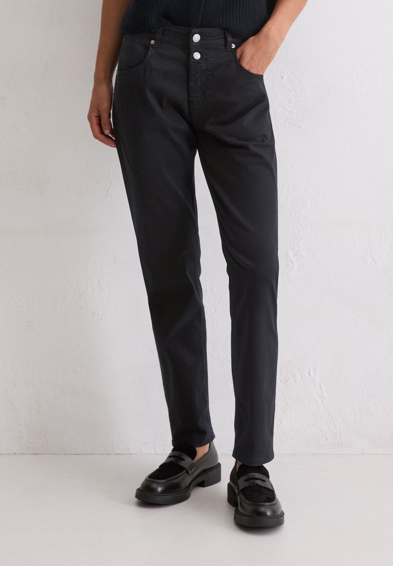 Personne portant un pantalon noir à jambes droites, des mocassins noirs et un haut sombre, une main dans la poche, devant un mur uni.