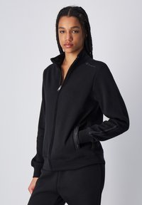 Sudadera negra con cremallera fabricada en un tejido suave, con cuello alto, bolsillos laterales y un logo en el hombro izquierdo. Detalles en costura contrastante.