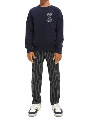 Kind trägt marineblauen Pullover mit weißem Logo, schwarz-grau gestreifte vertikale Hosen und weiße Turnschuhe mit marineblauen Akzenten.