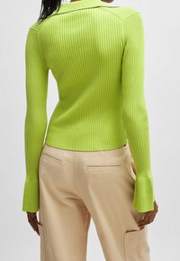 Dopasowany sweter prążkowany w jasnym limonkowym kolorze z kołnierzykiem i rozszerzającymi się mankietami, w połączeniu z beżowymi spodniami z bocznymi kieszeniami.