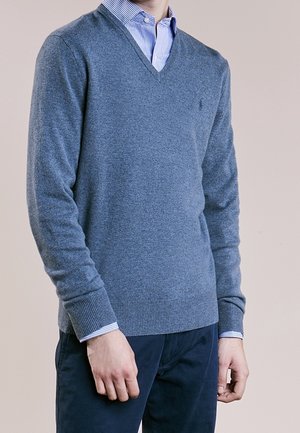 Homme portant un pull à col en V bleu par-dessus une chemise à carreaux bleue et un pantalon bleu foncé, debout devant un fond uni.