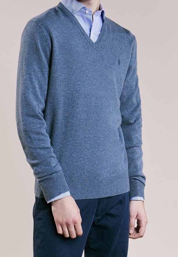 Homme portant un pull à col en V bleu par-dessus une chemise à carreaux bleue et un pantalon bleu foncé, debout devant un fond uni.