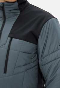 Giacca con colletto che presenta un corpo grigio medio e una sezione superiore nera. Il design include una texture trapuntata e una tasca sul petto con zip.