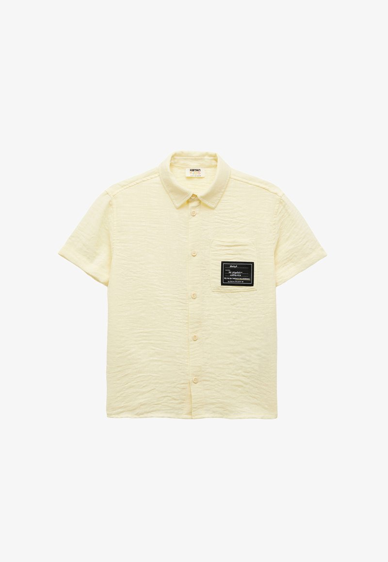 Jasnożółta koszula z krótkim rękawem o teksturowanej tkaninie, z zapięciem na guziki oraz czarną prostokątną kieszenią z białym napisem.