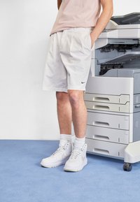 Bílé kraťasy ke kolenům s záhyby, malá černá Nike logo, v kombinaci s bílými vysokými teniskami a bílými ponožkami, stojící vedle kopírky.