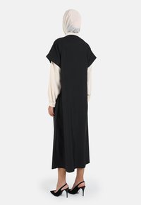 Black dress with short sleeves, layered over a cream blouse, featuring a straight cut and side slits. Accented with high-heeled shoes.