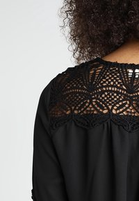 Czarna bluzka z misternym czarnym koronkowym wykończeniem przy dekolcie. Koronka ma kwiatowy i geometryczny wzór, co tworzy kontrast tekstur.