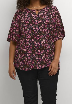 Person iført en sort kortærmet bluse med pink blomsterprint og sorte bukser, stående mod en ensfarvet lys baggrund.