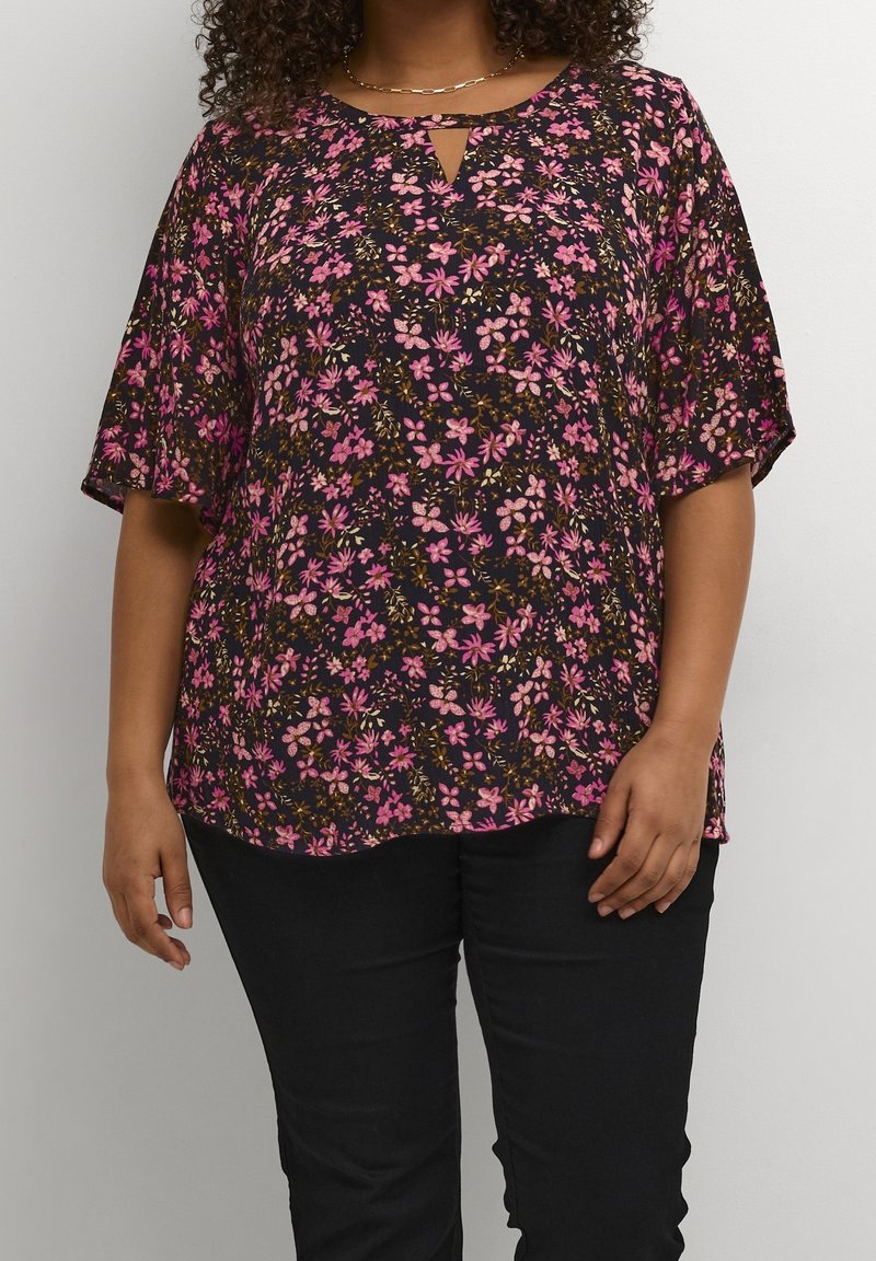 Personne portant un chemisier noir à manches courtes avec un imprimé floral rose et un pantalon noir, debout devant un fond clair uni.