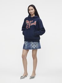 Jeune femme portant un sweat à capuche bleu marine avec le texte "Marc Jacobs", une jupe en jean bleue avec des patchs en forme de diamant, et des chaussures pointues argentées.