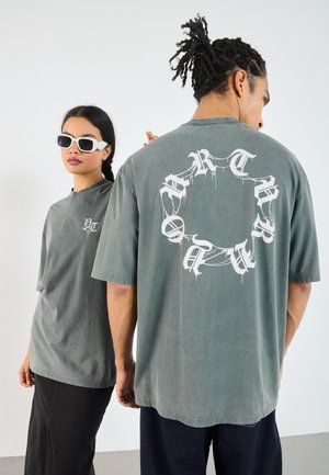Grå oversized t-shirts med grafiske designs foran og bagpå, korte ærmer og et blødt, struktureret stof. Kompletteret med hvide solbriller.