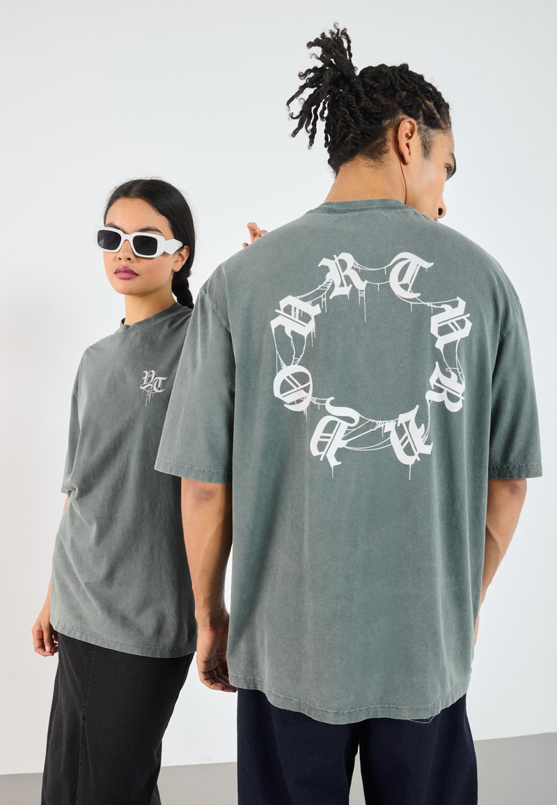 Grå oversized t-shirts med grafiske designs foran og bagpå, korte ærmer og et blødt, struktureret stof. Kompletteret med hvide solbriller.