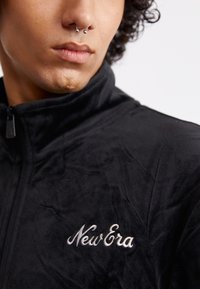 Sort fleecejakke med lynlås, med høj krave og hvid broderet "New Era" logo på brystet. Blød tekstur og glat finish.