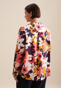 Camicia a fantasia floreale in colori vivaci, caratterizzata da una vestibilità rilassata, piega sul retro e orlo arrotondato. Il tessuto sembra liscio con maniche lunghe e dettagli con bottoni.