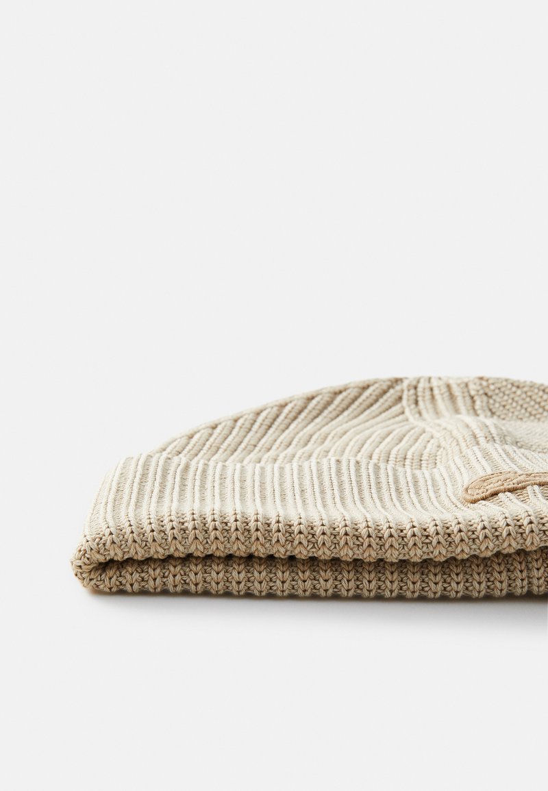 Beanie lavorato a maglia in beige, con costine testurizzate e bordo ripiegato, caratterizzato da un sottile dettaglio accentato su un lato.