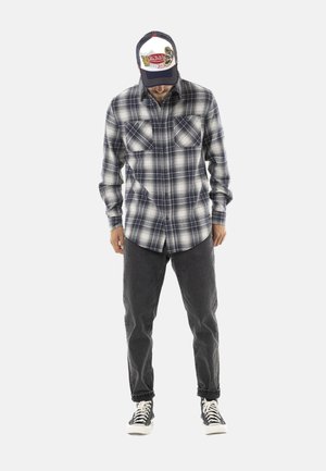 Homme portant une chemise à carreaux noire et blanche boutonnée, un jean noir, des baskets montantes noires et une casquette de baseball colorée avec un logo, regardant vers le bas.