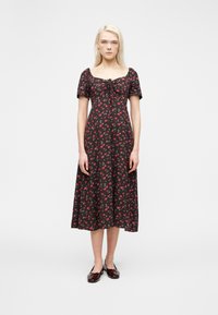 Γυναίκα με μακριά ξανθά μαλλιά που φοράει ένα μαύρο μίντι φόρεμα με κόκκινο λουλουδάτο σχέδιο, φουσκωτά μανίκια, λεπτομέρεια δέσιμου μπροστά και σκούρα καφέ άνετα παπούτσια.