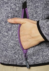 Szary sweter z dzianiny z fioletową kieszenią na zamek. Posiada czarną lamówkę na mankietach i u dołu. Ręka w kieszeni, pokazująca funkcjonalny design.