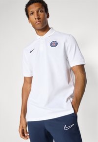 Hvid polo-shirt lavet af bomuld, med en knapkrave, sideslidser og et marineblåt Paris Saint-Germain-logo på venstre bryst.