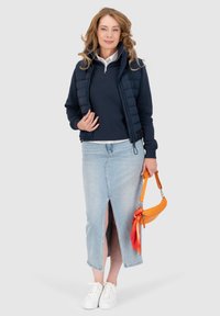 Gilet imbottito blu navy sopra un maglione blu navy, abbinato a una gonna in denim azzurro con uno spacco frontale. Sneakers bianche e una borsa arancione con una sciarpa.