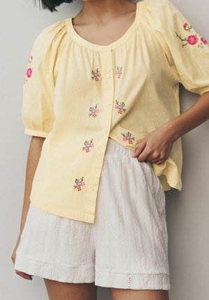 Kvinde iført en bleg gul knapbluse med lyserød blomsterbroderi og hvide teksturerede shorts, stående mod en ensfarvet baggrund.