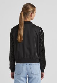 Schwarze Bomberjacke mit satiniertem Körper und paillettenbesetzten Ärmeln. Verfügt über gerippte Bündchen und Saum. Kombiniert mit hellblauen Jeans.
