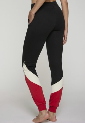 Black joggers with white and red chevron accents on lower legs, worn barefoot, with a zippered side pocket visible on the right.