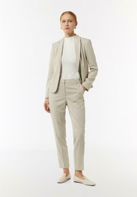 Jasno beżowy garnitur szyty na miarę z dopasowaną marynarką i wąskimi spodniami, w połączeniu z białym golfem i beżowymi butami wsuwanymi. Gładka faktura, minimalne detale.