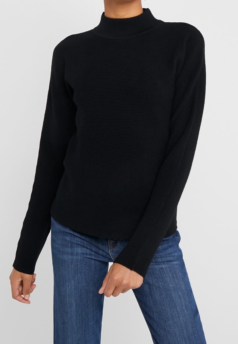 Svart ribbad polotröja med långa ärmar, strukturerat tyg och avslappnad passform. Kombinerad med blå jeans, händerna placerade vid midjan.