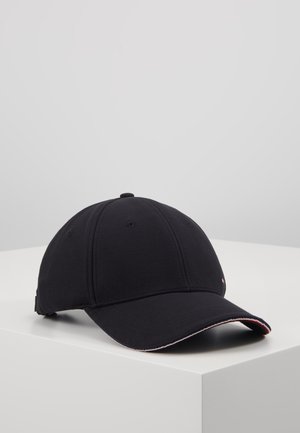 Czarna czapka z daszkiem z zakrzywionym rondem i małym czerwono-biało-niebieskim detalem na krawędzi daszka, umieszczona na białej powierzchni.