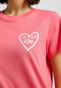 T-shirt en coton rose avec un design de cœur blanc portant le texte "Lee" et les chiffres "1889". Manches courtes et col rond.