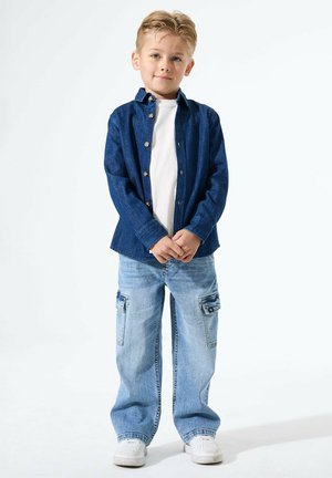 Blauwe denim-overhemd met knopen, witte t-shirt eronder, lichtblauwe wijde jeans met zakken en witte sneakers.